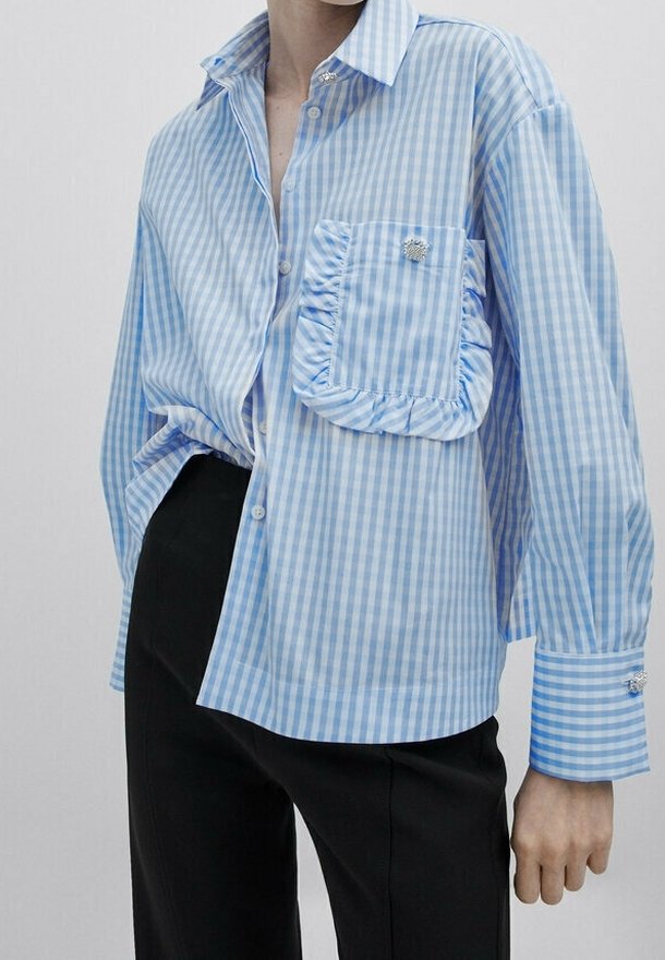 Blau-weiß gestreifte Button-Down-Bluse mit einer gefältelten Tasche, langen Ärmeln und silbernen Knopfakzenten. Getragen mit schwarzen Hosen.