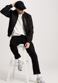 Zwarte bomberjack over een wit shirt, gecombineerd met zwarte jeans. De schoenen zijn grijs met witte accenten. Het model zit op een witte kruk.