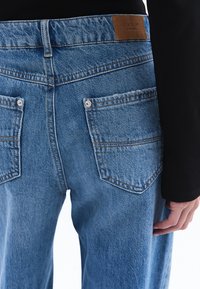 Vaaleansiniset farkut, joissa on korkea vyötärö, sisältävät kaksi takataskua metallinitteillä ja ruskean nahkapatchin vyötärönauhassa.