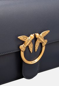 Borsa a tracolla in pelle blu navy con una chiusura ad anello dorato che presenta due uccelli stilizzati. Texture liscia con accenti di cucitura visibili e chiusura a pattina.