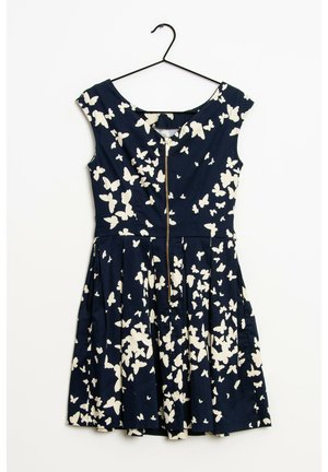 Robe bleu marine sans manches avec des motifs de papillons blancs et une fermeture éclair à l’avant, suspendue sur un cintre noir sur fond blanc.