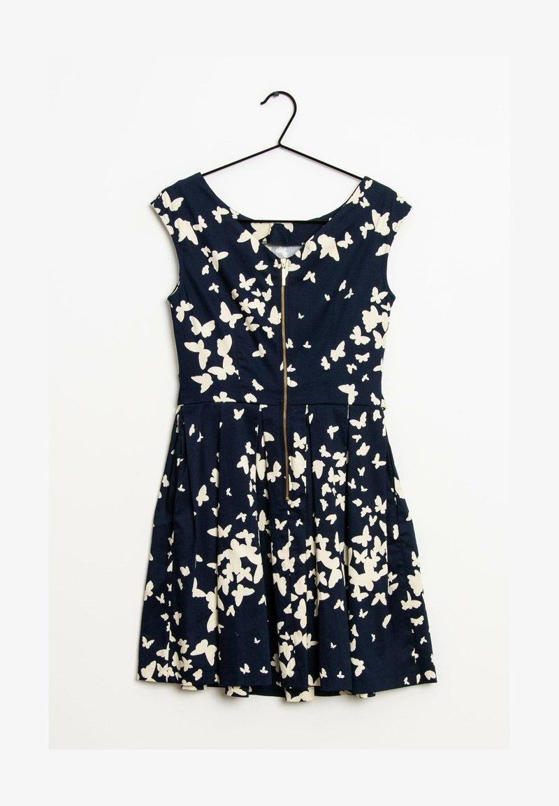 Robe bleu marine sans manches avec des motifs de papillons blancs et une fermeture éclair à l’avant, suspendue sur un cintre noir sur fond blanc.