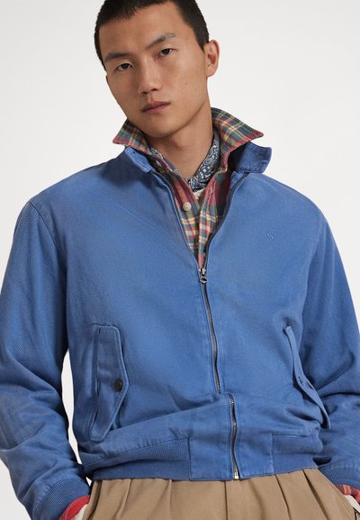 Μπλε ζεκακι με φερμουάρ και ψηλό γιακά, με πλαϊνές τσέπες και ριμπ μανσέτες. Φορεμένο πάνω από καρό πουκάμισο και με μαντίλι με σχέδια.