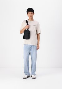 Svart crossover-veske med glidelåslomme, laget av slitesterkt materiale og justerbar stropp. Bæres over en beige skjorte og lyseblå jeans.