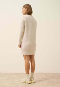 Sweterkowa sukienka z długim rękawem w odcieniu kości słoniowej z teksturowym wzorem. Połączona z kremowymi sneakersami i skarpetkami, wyeksponowana na jasnym tle.