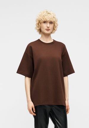 Woman with short curly blonde hair wearing a loose brown short-sleeve shirt and black pants, standing against a plain light background.