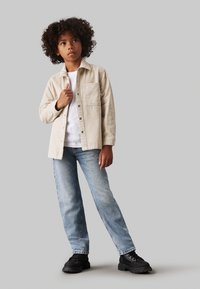 Camicia in corduroy beige con tasca frontale, t-shirt bianca sotto, jeans blu chiaro a gamba dritta e stivali neri chunky.