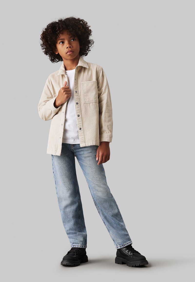 Camicia in corduroy beige con tasca frontale, t-shirt bianca sotto, jeans blu chiaro a gamba dritta e stivali neri chunky.