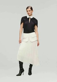 Bodysuit noir avec un col haut et des détails de cape, associé à une jupe plissée blanche et des bottes noires à talons montantes.