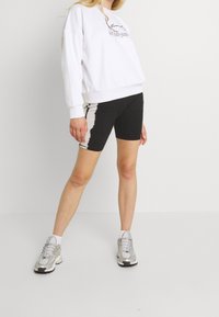 Sudadera blanca con logotipo, pantalones cortos de bicicleta negros con patrón de tie-dye y zapatillas plateadas. Tejido suave, corte relajado y estilo atlético.