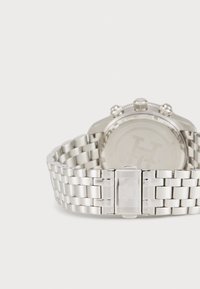 Silberne Armbanduhr mit einem Armband aus rostfreiem Stahl, runder Gehäuse und einem Verschluss mit einem gravierten Logo. Das Ziffernblatt der Uhr ist minimalistisch und glatt.