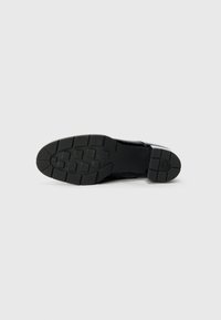 Suela de zapato de goma negra con un patrón de agarre texturizado que presenta formas cuadradas y rectangulares, detalles en el talón y un perfil plano.
