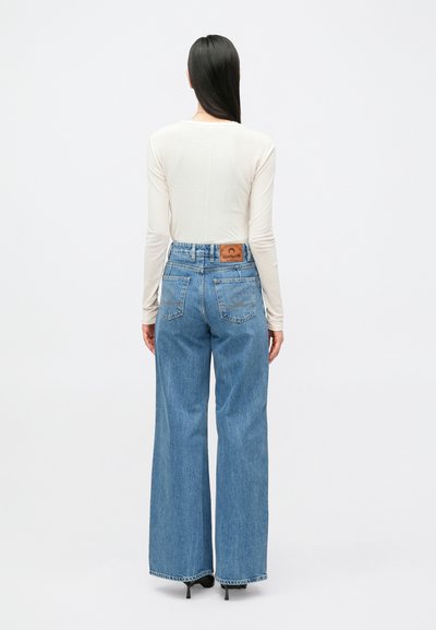 Φως μπεζ μπλούζα με μακριά μανίκια συνδυασμένη με ψηλόμεσα, ανοιχτό μπλε, παντελόνια με φαρδιά πόδια που έχουν καφέ δερμάτινο ταμπελάκι και κλασικές πίσω τσέπες.