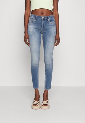 Persoon die lichtblauwe skinny jeans draagt, een cropped lichtgroene top en beige sandalen met bandjes, staand tegen een effen lichte achtergrond.
