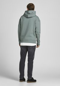 Mikina s kapucí v tlumené zelené barvě, z bavlněné směsi, s volným střihem a žebrovaným lemem. Nosič ji má spolu s tmavě šedými džíny a černými teniskami.