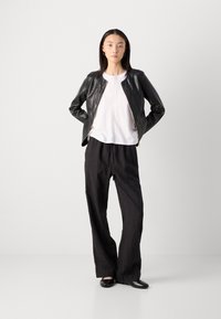 Zwarte leren jack met ritssluiting, witte top en losse zwarte broek. Model draagt zwarte flats en zilveren hoepeloorbellen. Eenvoudig, modern ontwerp.