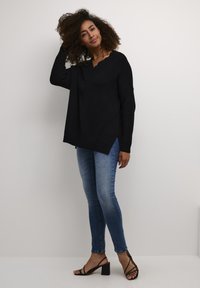 Schwarzer, gestrickter Oversized-Pullover mit V-Ausschnitt und seitlichen Schlitzen, kombiniert mit blauen Slim-Fit-Jeans und schwarzen, hohen Sandalen.