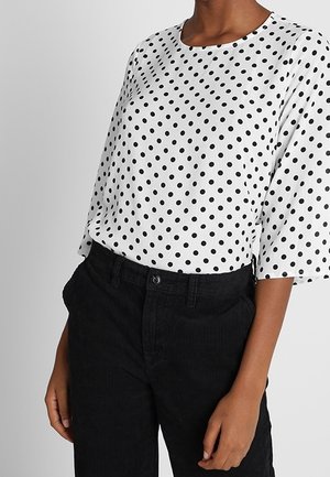 Valkoinen pusero mustilla pilkulla, löysä malli, pyöreä kaula-aukko ja lyhyet hihat, yhdistettynä mustiin samettipö trousers, joissa on taskut.