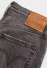 Pantalones vaqueros de mezclilla gris con un parche de cuero que presenta el logotipo de la marca, diseño de cinco bolsillos y costuras en contraste. La tela parece suave y desgastada.