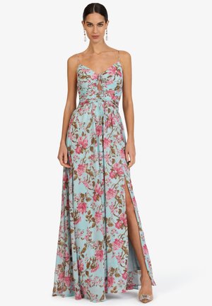 Frau trägt ein ärmelloses, hellblaues Maxikleid mit floralem Muster, Frontschlitz und dünnen Schulterriemen, steht mit einem freigelegten Bein.