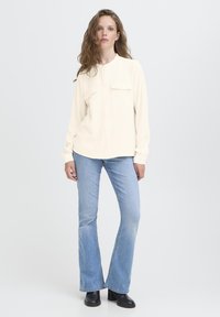 Kvinde med langt bølget hår iført en cremegul langærmet skjorte med lommer, lyseblå flared jeans og sorte hælede støvler, stående på hvid baggrund.