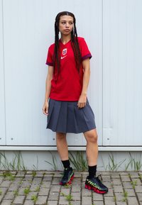 Red vertical-striped athletic shirt with a crest; gray pleated skirt; black shoes with green and red accents; worn with black socks.