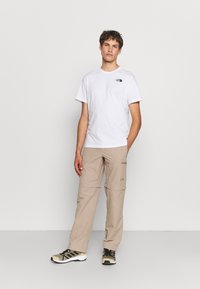 Camiseta blanca de algodón con un pequeño logo negro, combinada con pantalones cargo beige que tienen bolsillos y cremalleras, usados con zapatos negros y beige.
