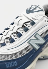 Detailní pohled na bílou a modrou tenisku New Balance 1000, která ukazuje síťovinu, tkaničky a logo "N" po straně.