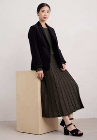 Blazer di velluto nero indossato sopra un abito scuro a pieghe con finitura metallizzata. Sandali con plateau neri, in piedi accanto a un blocco di legno chiaro.