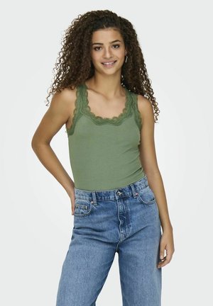 Lichtgroene geribbelde tanktop met kantafwerking, gecombineerd met hoge taille blauwe spijkerbroek. Model poseert met hand op de heup tegen een effen achtergrond.