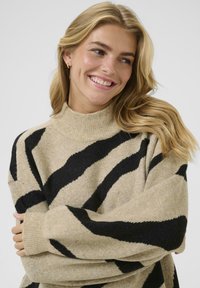 Beige sweater met zwarte zigzagstrepen, hoge hals, geribd textuur en brede mouwen. Zacht materiaal met een ontspannen pasvorm.