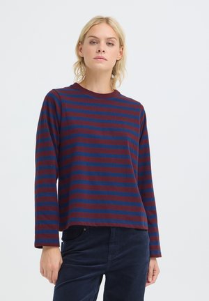 Femme aux cheveux blonds portant un chemisier à manches longues rayé bordeaux et bleu marine et un pantalon sombre sur un fond blanc uni.