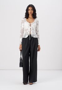 White floral lace top with long sleeves and a tie-front detail, paired with loose black trousers. Model holds a small black handbag.