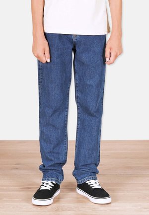Siniset denim-farkut suoralinjaisella leikkauksella, joissa on klassinen viiden taskun muotoilu ja hienovaraiset ompeluyksityiskohdat, yhdistettyinä mustiin tennareihin.