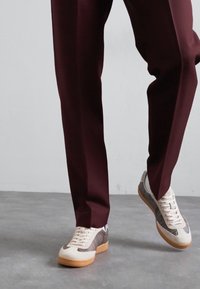 Sneakers bianchi con accenti in rete grigia, suola in gomma beige, abbinati a pantaloni su misura bordeaux, che mostrano una texture liscia e un design elegante.