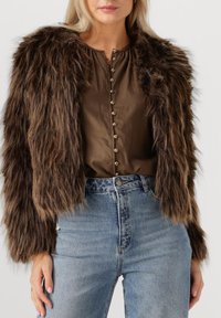 Nagesimuleerde bont cropped jacket in donkerbruin, met een harige textuur. Gdraagd over een bruine stoffen top met knopen en gecombineerd met lichtblauwe jeans.