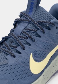 Blå löparskor med meshöversida, texturerade detaljer, bruna snörningar och en framträdande gul Nike Swoosh-logotyp på sidan.