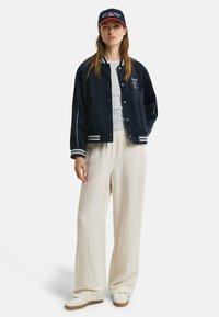 Marineblauer Bomberjacke mit weißen Streifenakzenten, grauer Rundhals-T-Shirt, creamfarbene weite Hose und weiße Sneakers mit brauner Sohle; rot akzentuierte Mütze.