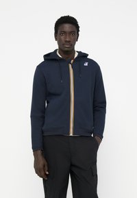 Marineblå zip-up hættetrøje lavet af bomuldsblanding, med en stribet lynlås, justerbar hætte og ribbede manchetter. Logo-patch på brystet.
