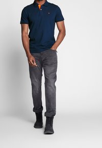 Polo shirt navy avec des accents de col orange, associé à un jean gris et des bottes noires. Le matériau semble être du coton, avec une coupe classique et des manches courtes.