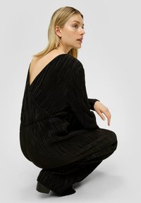 Blonde vrouw in hurkende positie, gekleed in een lange zwarte geplooide jurk met een diepe V-rug, kijkt naar rechts tegen een effen grijze achtergrond.