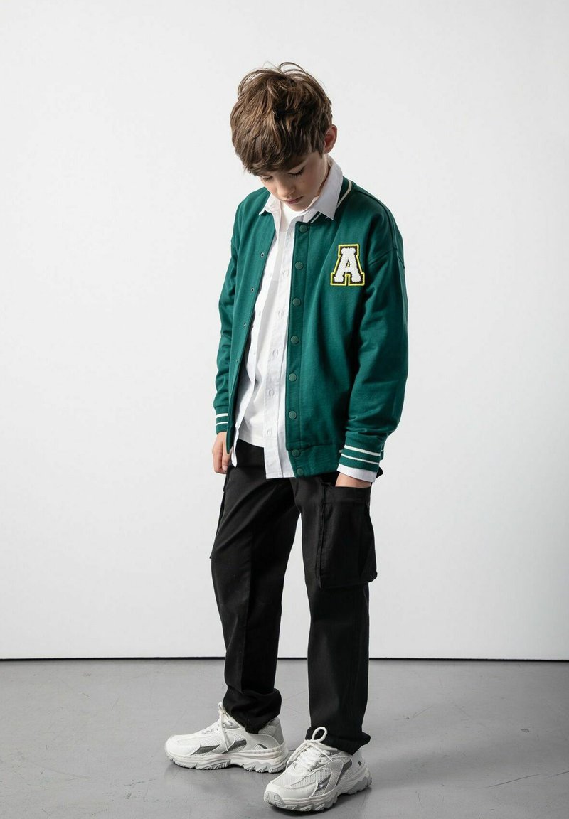 Zielona kurtka varsity z białymi paskami przy mankietach, z haftowaną literą "A". Połączona z białą koszulą i czarnymi spodniami cargo. Białe sneakersy.