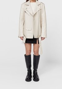 Weißes Leder-Bikerjacke mit diagonalen Reißverschlüssen, Fronttaschen und einem Gürtel-Detail. Getragen über einem cremefarbenen Pullover und einem schwarzen Minirock mit knieb hohen schwarzen Stiefeln.