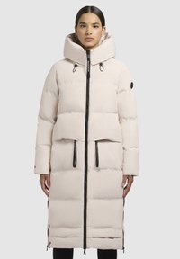 Woman wearing a long, light beige padded coat with large hood and black zipper details, standing against a plain background.