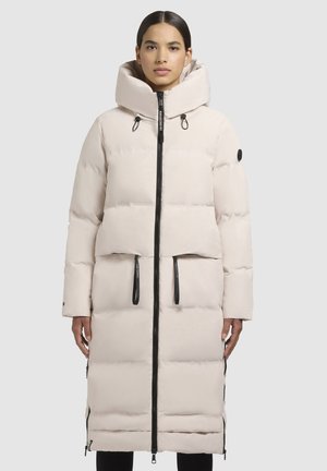 Vrouw in een lange, lichtbeige gewatteerde jas met een grote capuchon en zwarte ritsdetails, staand tegen een effen achtergrond.