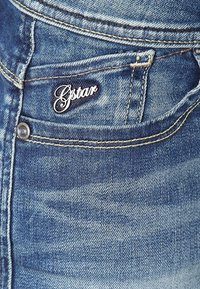 Blå denimjeans med en urtvättad textur, med en G-star-logotyp på framfickan och synlig söm längs sömmarna.
