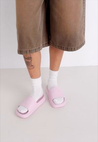 Lyserøde gummisandaler med åben tå, prydet med præget logo, iført med hvide sokker og brune shorts i knæ-længde.