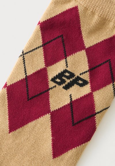 Tissu tricoté avec motif losanges argyle beige et rouge foncé, présentant des losanges noirs en contour et des lettres noires "BP" au centre.