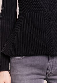 Černý žebrovaný pletený svetr s texturovanými vzory a peplum střihem, zkombinovaný se svetle šedými džíny, které mají hladký povrch.