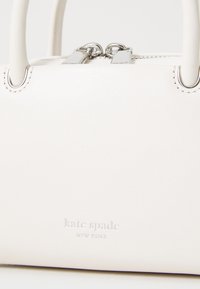 White leather handbag with silver hardware, featuring two top handles, zip closure, and embossed logo on the front. Compact design.